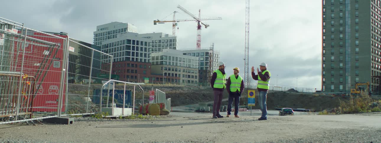 Three construction workers are standing outside talking near a construction site with buildings and a crane