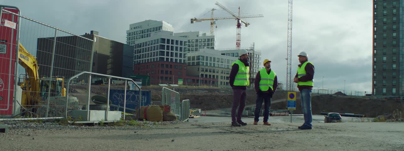 Three construction workers are standing outside talking and one man is pointing to something and there are buildings in the distance