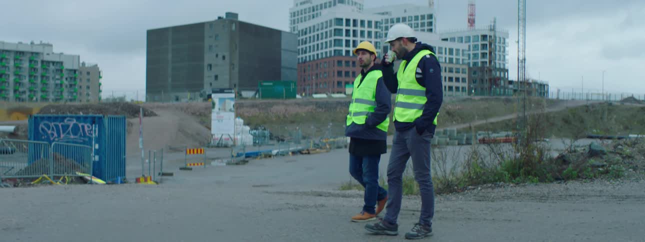 Two construction workers are walking outside and one is holding a cup as they talk