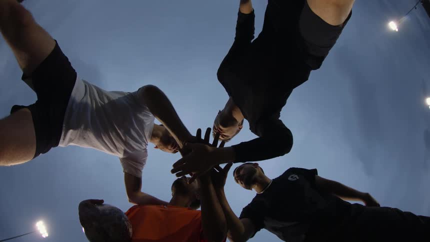 A group of men place their hands together while in a huddle outdoors in the evening