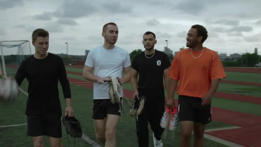 Four friends walking across a track toward the soccer field carrying their cleats and a ball