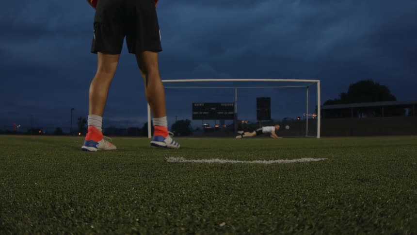 A player takes a free kick and scores against a leaping goalie on a soccer field