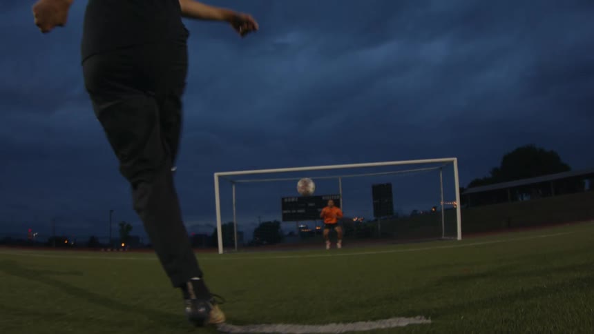 A soccer player kicks the ball towards the goal and it is blocked by the goalie