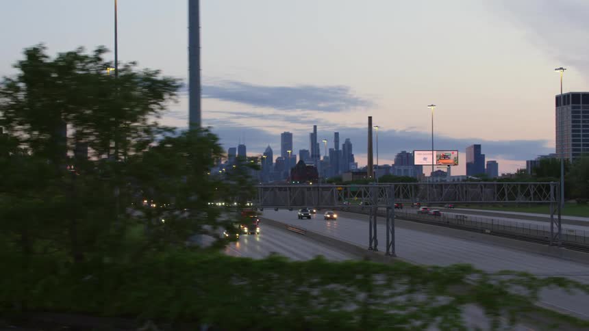 Vehicles travel along a highway in the evening with the city skyline behind