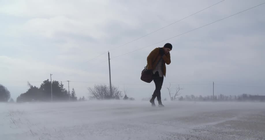 A man is walking along a field with snow blowing around him