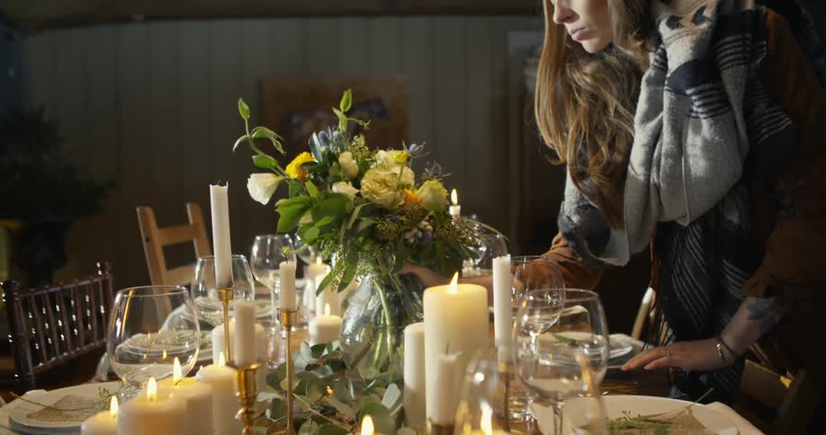 A woman places a vase of flowers in the center of a set dinner table