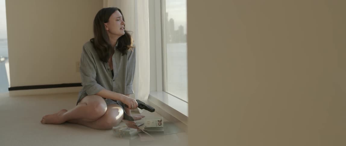 A woman sitting on the floor next to a window while she cries and holds a gun