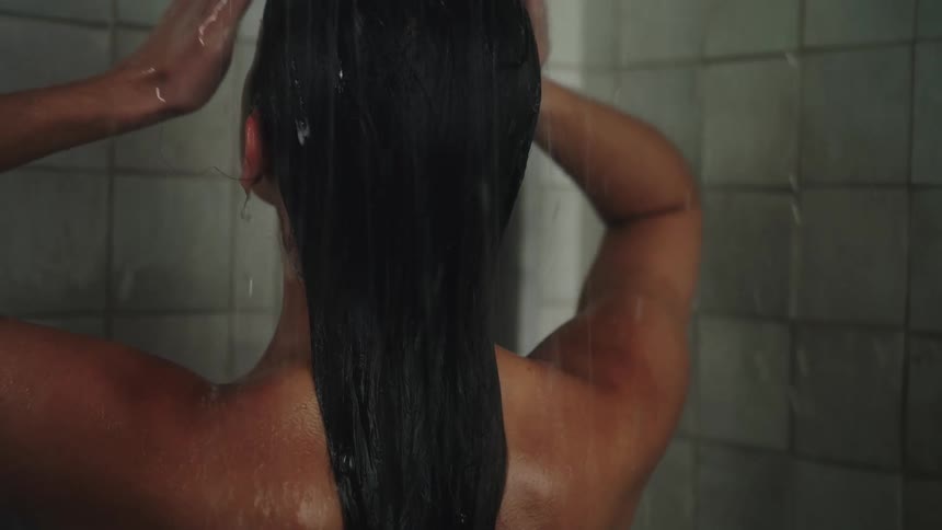 A woman is standing in the shower and moving her hands down her long dark hair as water falls on it