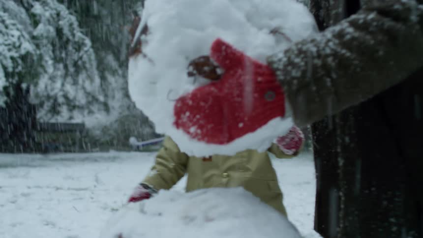 A parent helps a little boy carry a large snow ball and place it on their snowman