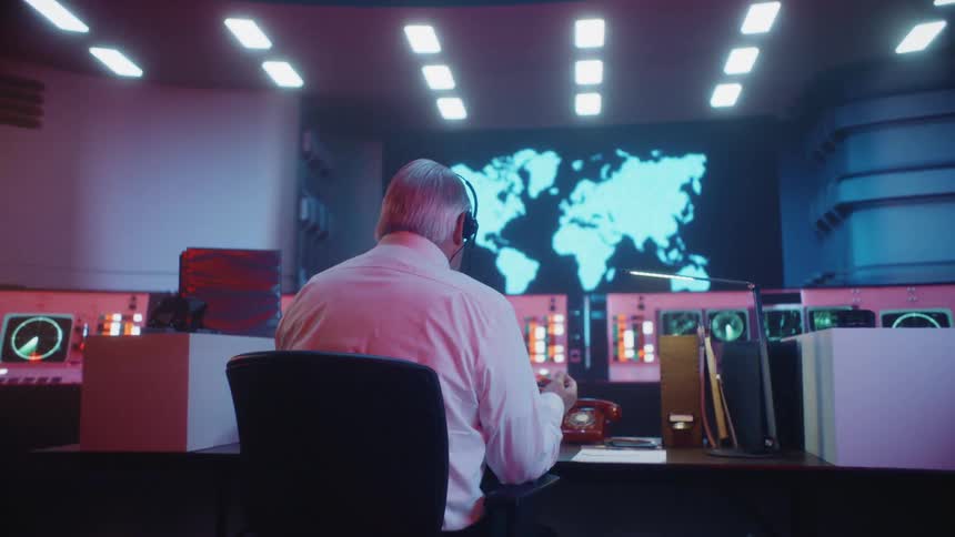 A man sitting at a desk in a control room gestures as he talk on his headset
