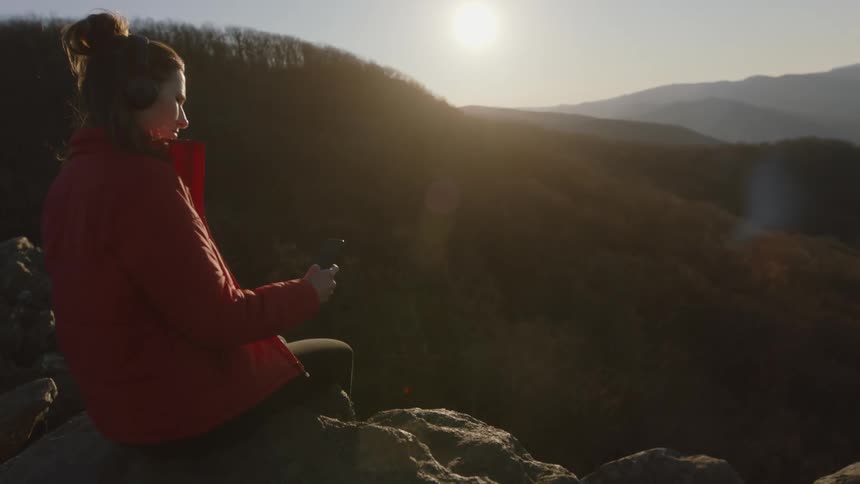 A woman wears headphones, enjoys music, and looks out over mountain view
