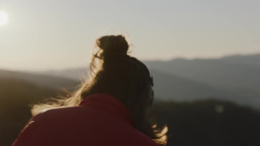 White woman sits in the golden glow of sunset and looks out over blue ridge mountains while listening to music