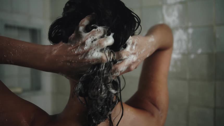 A woman is standing in the shower and there are soap bubbles on her hands as she washes her long dark hair