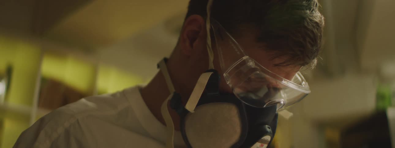 A man wearing safety glasses and a respirator mask looks down while working with his hands