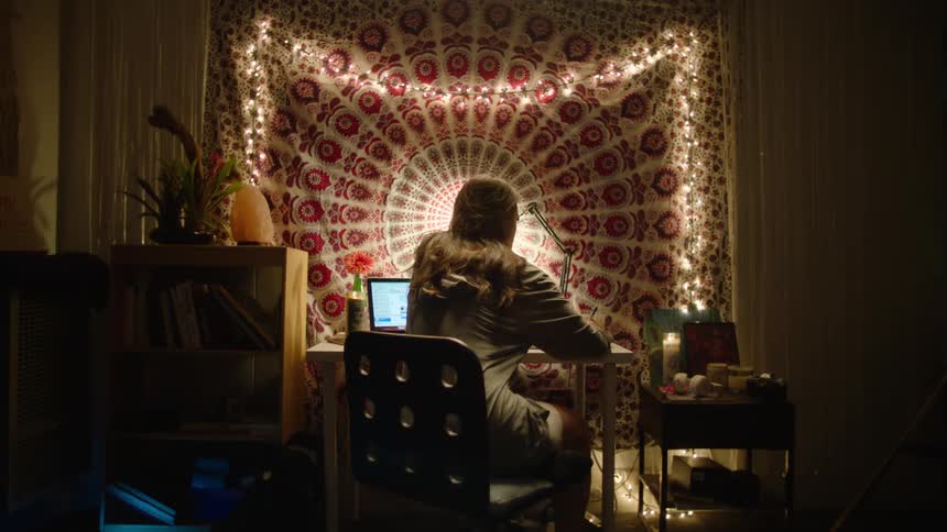A woman sitting at her desk writing with her laptop open and a decorative fabric covering the window