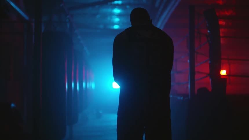 A man swings his arms while standing in a dimly lit boxing gym during the day
