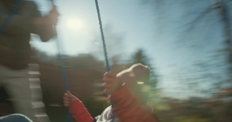 Girl with white wooly hat and grat coat pushes girl on swing in playground