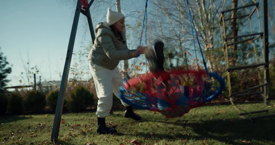Mother swings child sitting on hoop and mesh swing back and forth in backyard of home