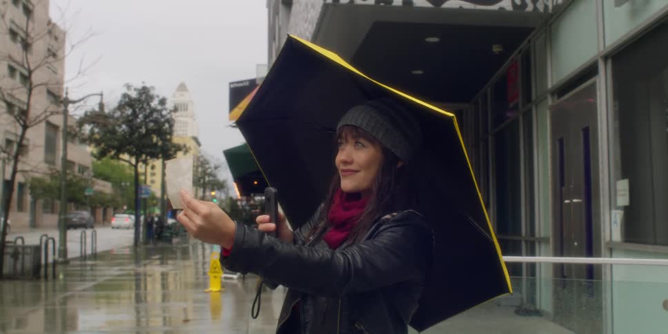A woman with an umbrella is standing on sidewalk near a street holding a photograph and her phone with a big smile on her face