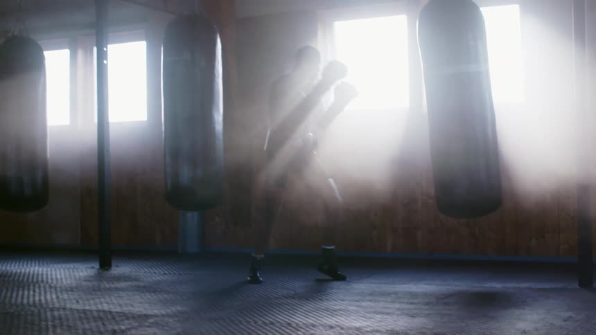 Male boxer with black trunks boxing gloves and shoes hits punching ball while the sun shines through windows