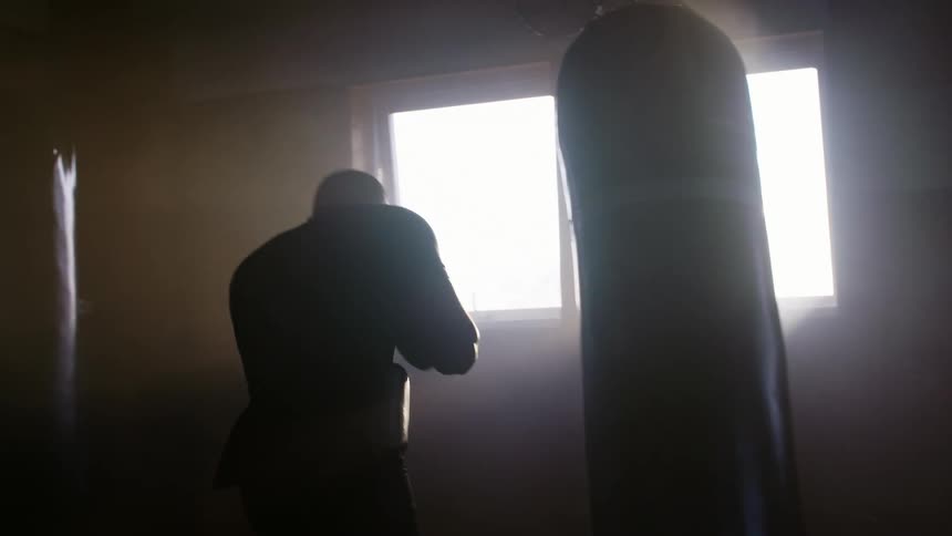A boxer trains by striking a punching bag in a dimly lit room during the day