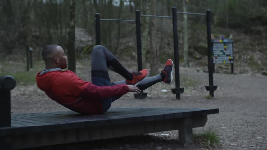 Young man wearing a red jacket lays over a wooden deck in a public park exercising