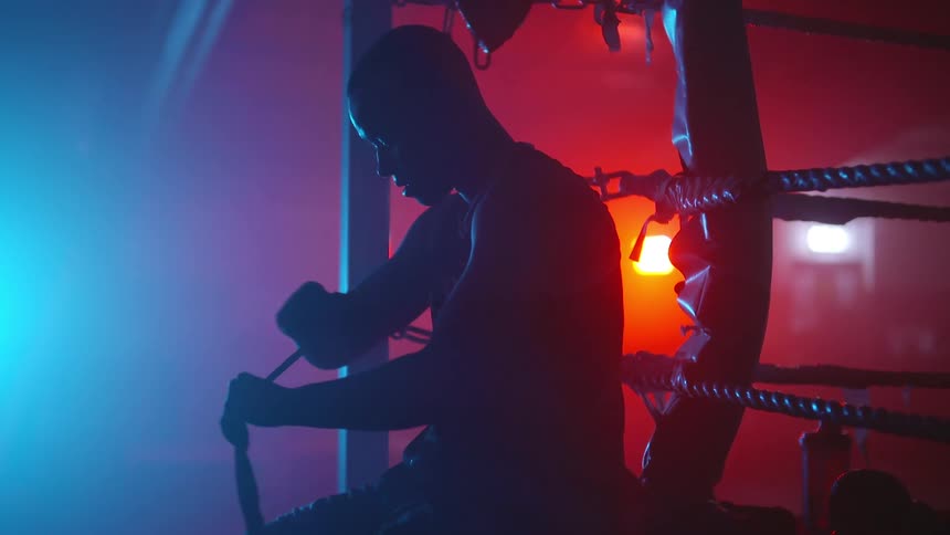A man is sitting on the edge of a boxing ring wrapping tape around his wrist