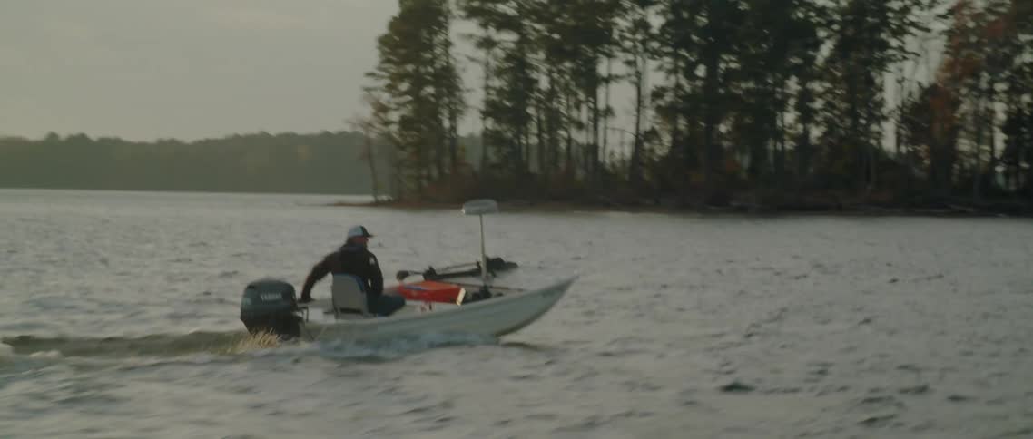 A man is sailing in a small motor boat near land with trees