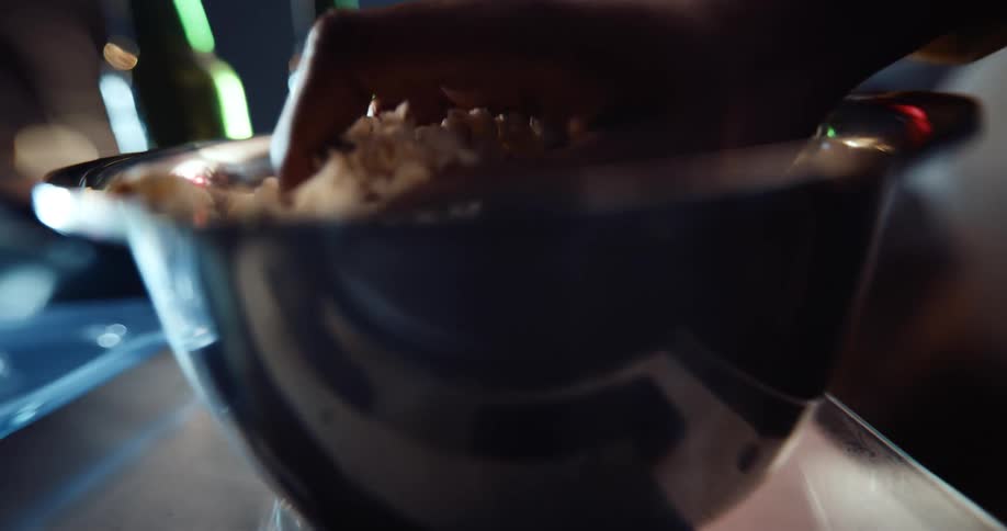 Person sits eating popcorn from metal bowl sitting next to two glass beverage bottles