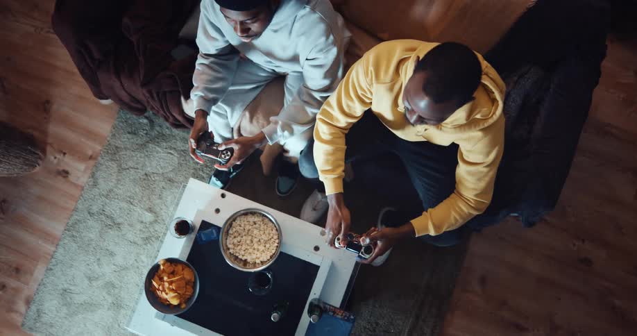 Two men are sitting on a couch playing video games and they have bowls of food on the table