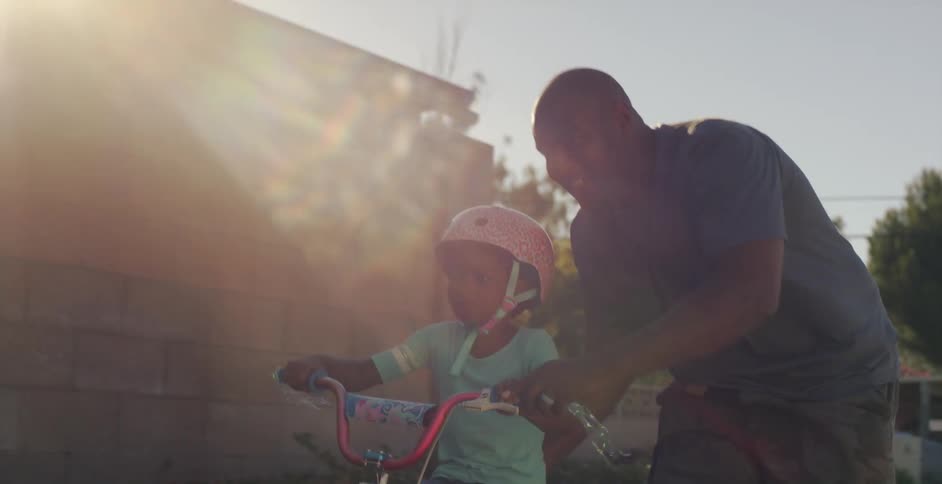A father smiles as he pushes his daughter down a neighborhood street on a two wheeled bicyle