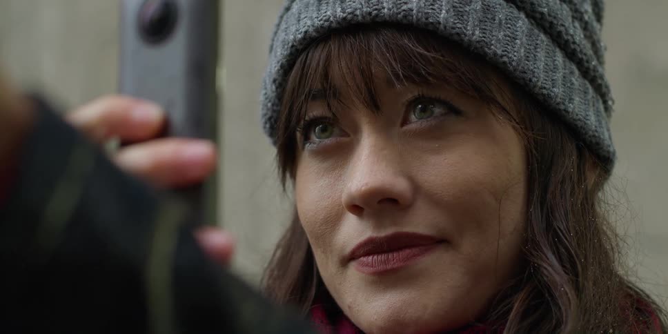 An asian woman wearing a gray knit cap is outside holding a camera phone is getting ready to take a picture
