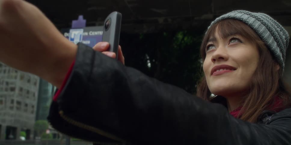 An asian woman wearing a gray knit cap is standing on a street holding a camera phone with a big smile on her face