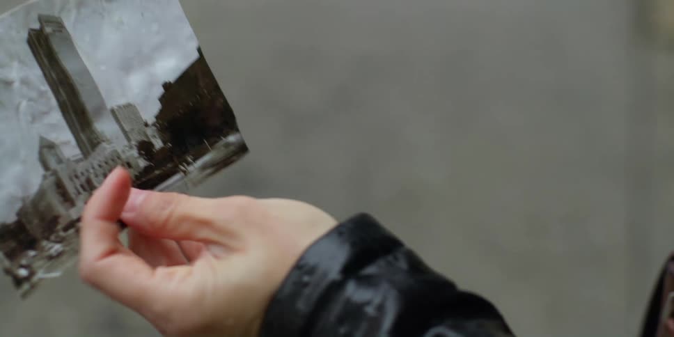 A woman holds a photograph of a building and takes out a camera to take a picture of it