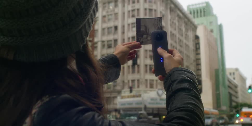 A woman holds a black and white photo up against a downtown building and takes a picture