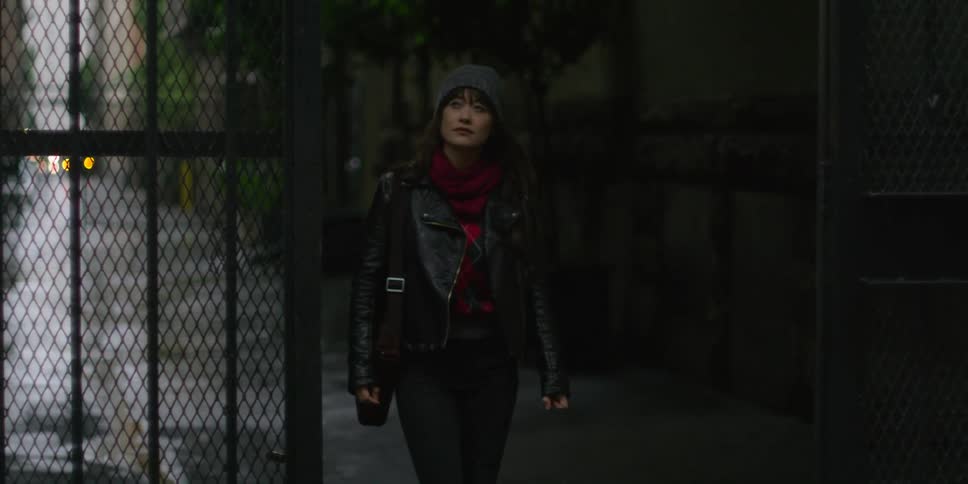 A woman wearing a grey beanie and black leather jacket and brown leather bag walks down narrow street in the city