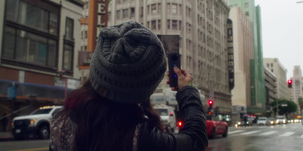 A woman wearing a gray knit cap is standing on a downtown street holding a photograph and a camera