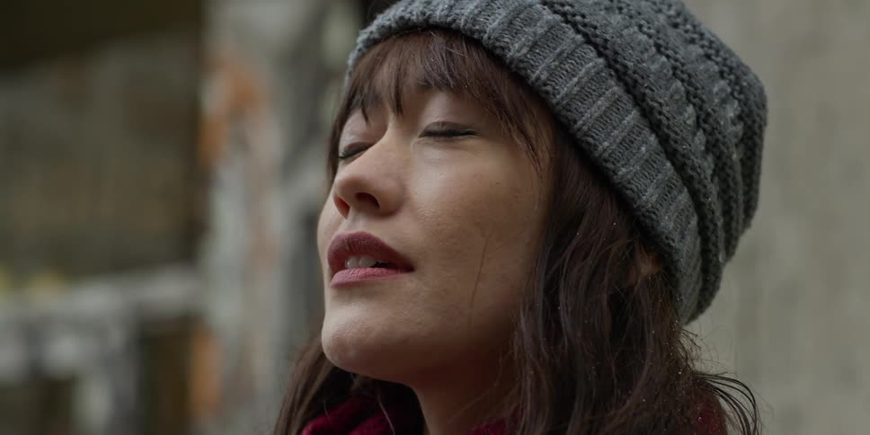 An asian woman wearing a gray knit cap is outside looking up