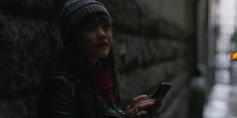 Woman wearing a grey beanie and black leather jacket leans against the wall using smartphone in narrow street