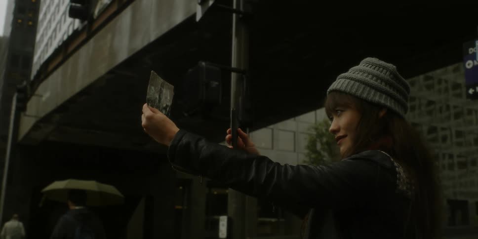 A woman is standing on a city sidewalk using her phone to take a picture of a photograph