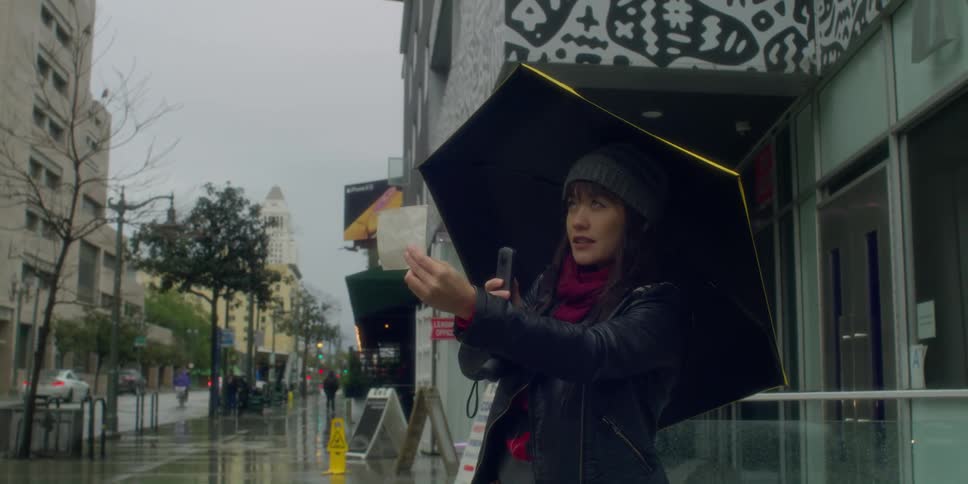 A woman holding up a photograph taking a picture with her phone while standing on a city sidewalk in the rain
