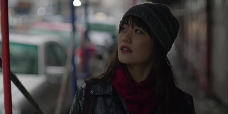 An asian woman wearing a grey knit cap is walking near buildings on a busy street as she looks around