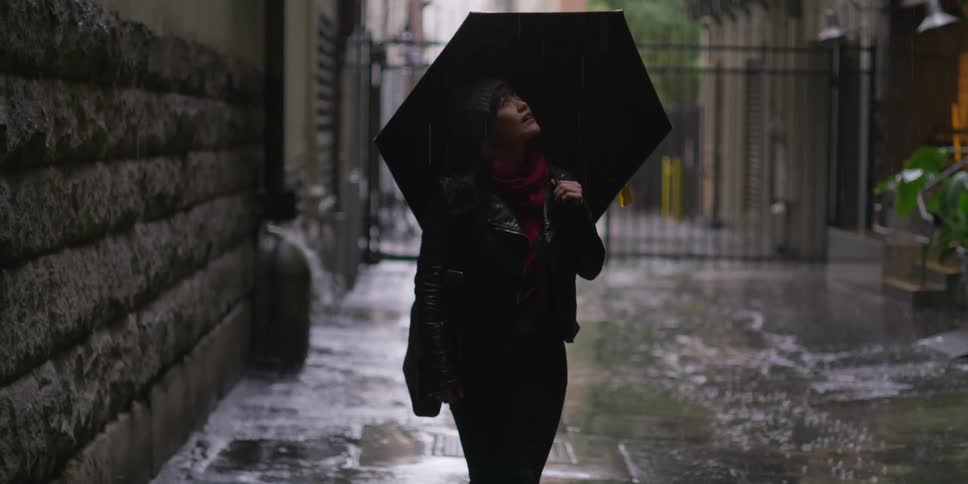 A woman is outside walking in the rain with an umbrella looking up and she stops near a building and takes down her umbrella