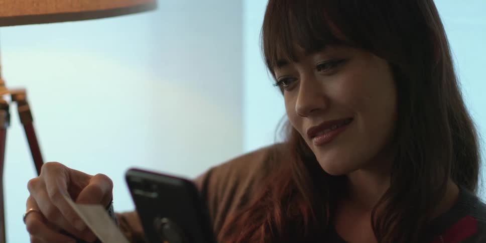 A woman sits and smiles at a photograph then lowers it and looks at her phone