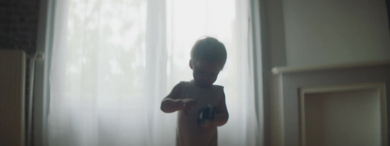 A toddler plays with a toy car while standing in front of a curtained window indoors during the day