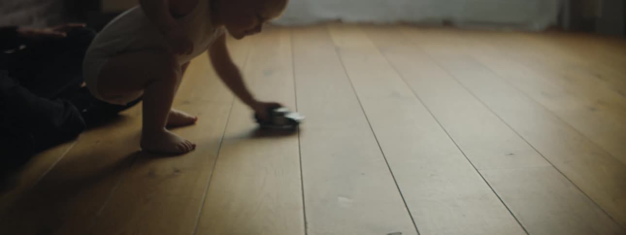 Toddler boy wearing a t shirt and a diaper crouches down playing with toy car in the sunlight coming through the curtains on the window stumbles and the car slips out of his hand