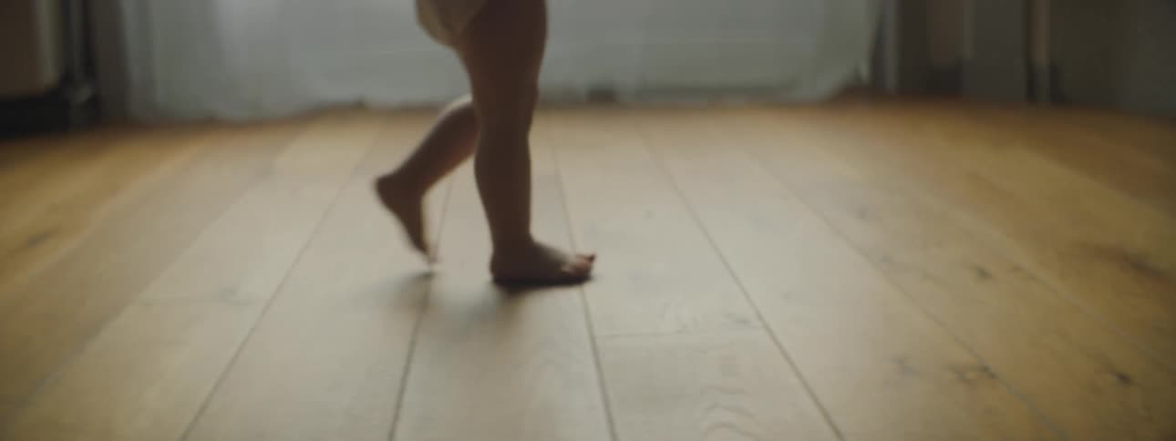 A diapered baby walking barefoot across wood floors inside a home