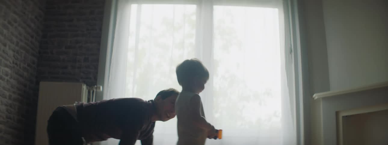 Father smiles on all fours as toddler walks away carrying plastic toy
