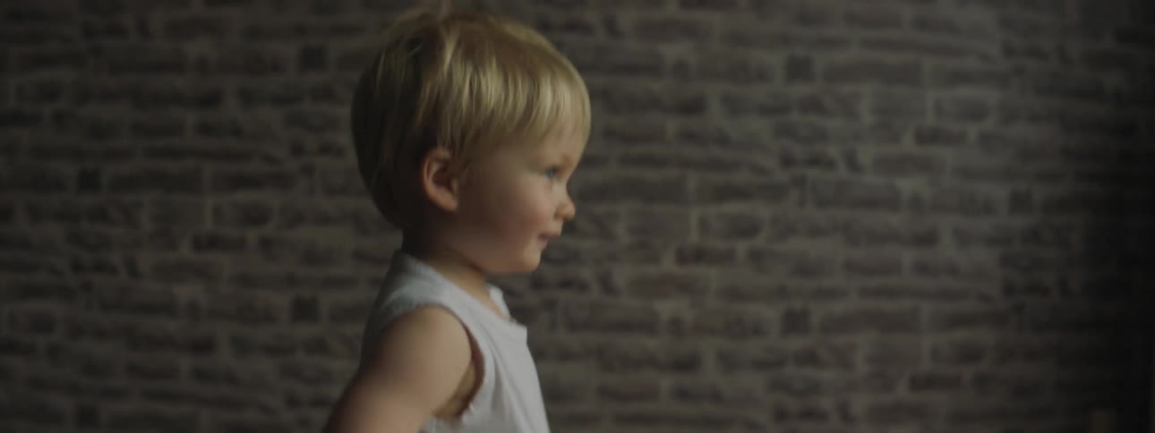 Blonde toddler walks out of door past brick wall to accept wooden toy from person