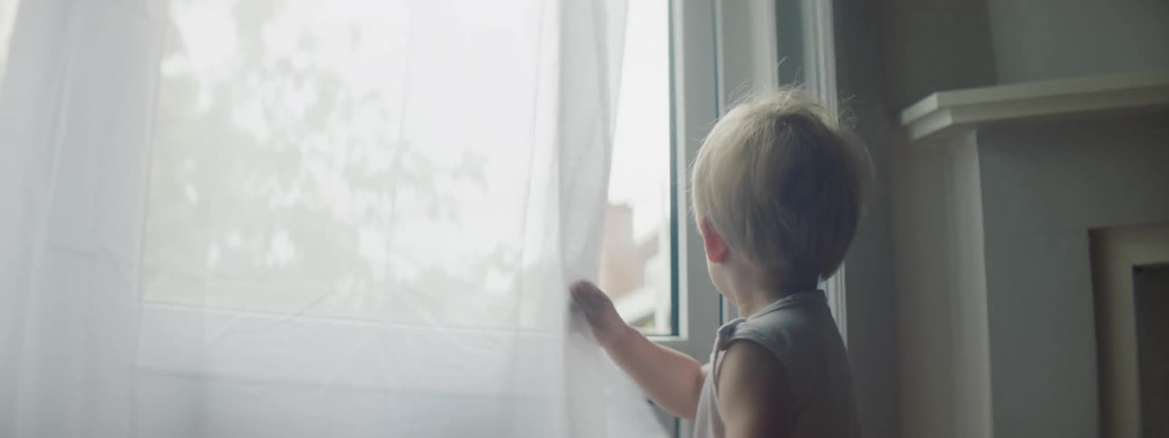 A young toddler boy grabs the sheer curtains and walks behind it